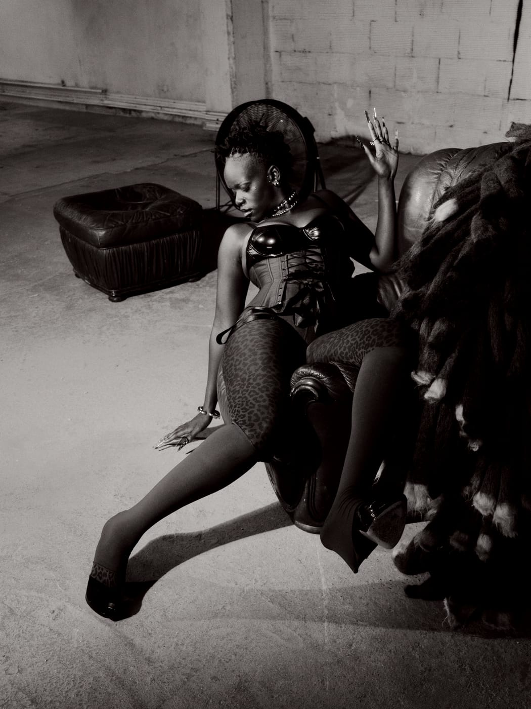 Ezek seated at the armchair's edge, one hand raised, leopard tights, toned monochrome