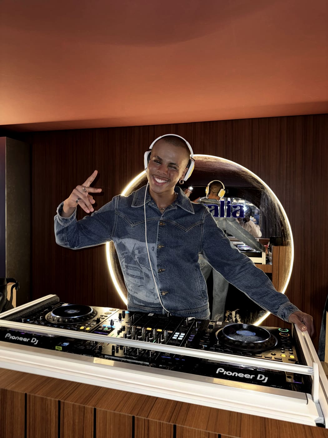 Oluwa in a denim jacket behind the decks, headphones on, two fingers raised